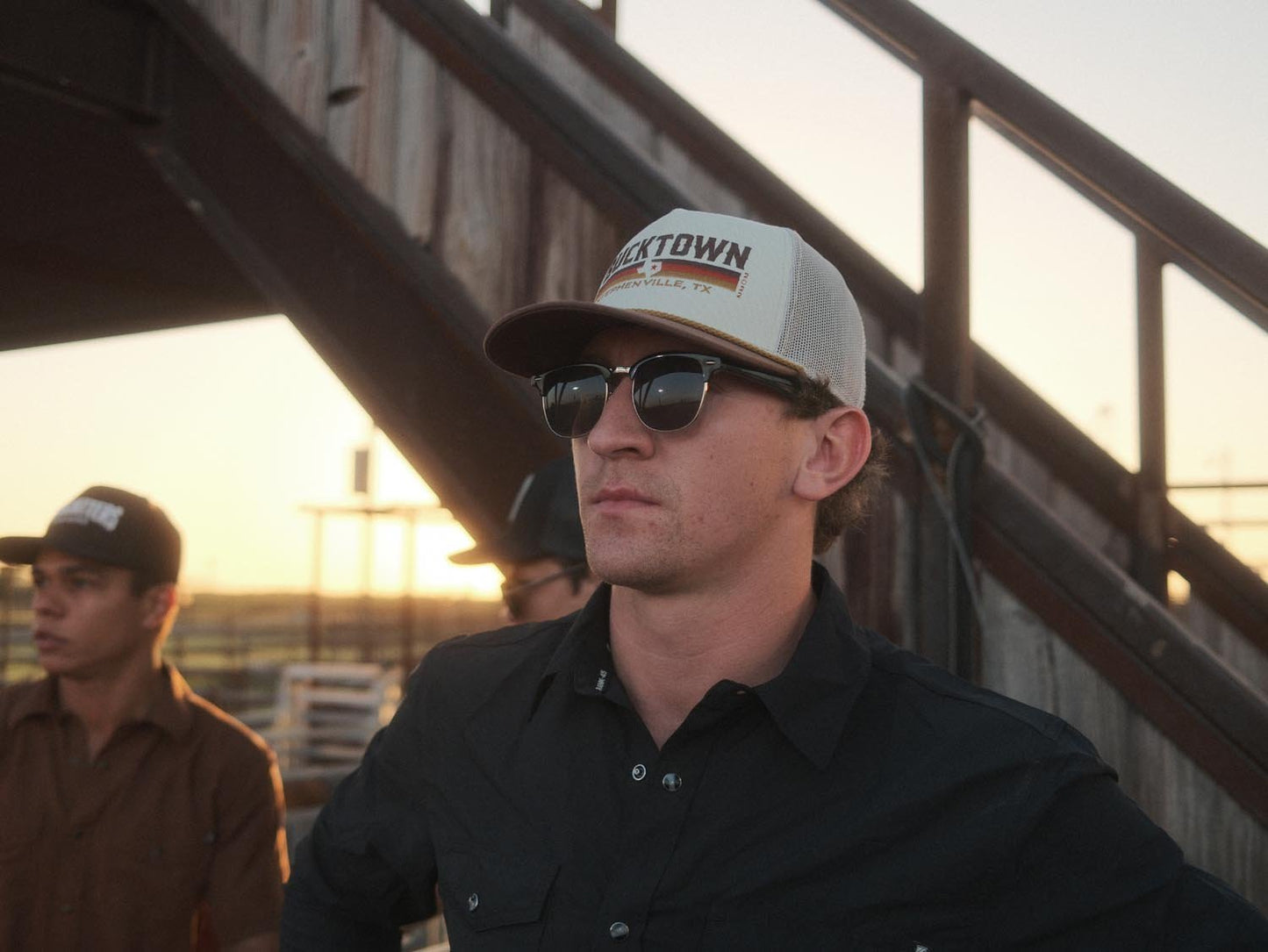 A man in sunglasses, a black shirt, and a Texas star hat stands by a wooden staircase at sunset. Another man in the background wears the Bucktown Revival Hat by Rural Cloth.