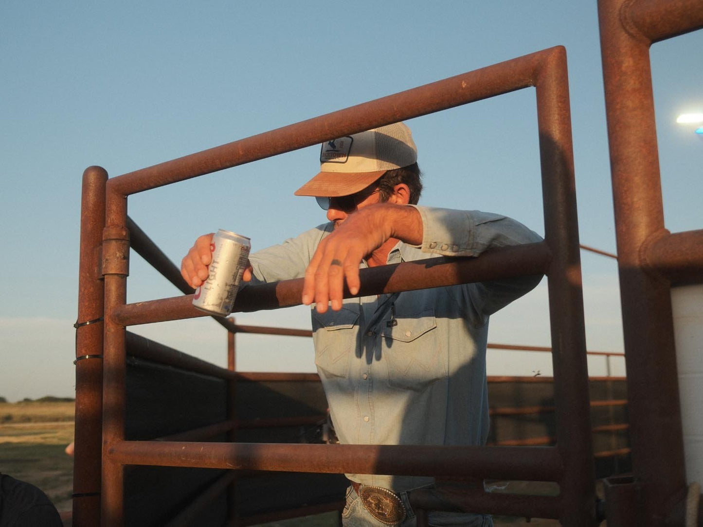 Wearing the Rural Cloth Last Out Hat, sunglasses, and a light blue shirt, a person leans on a rusty gate with a can in hand. An open field stretches out under a clear sky at Bucktown XV Ranch.