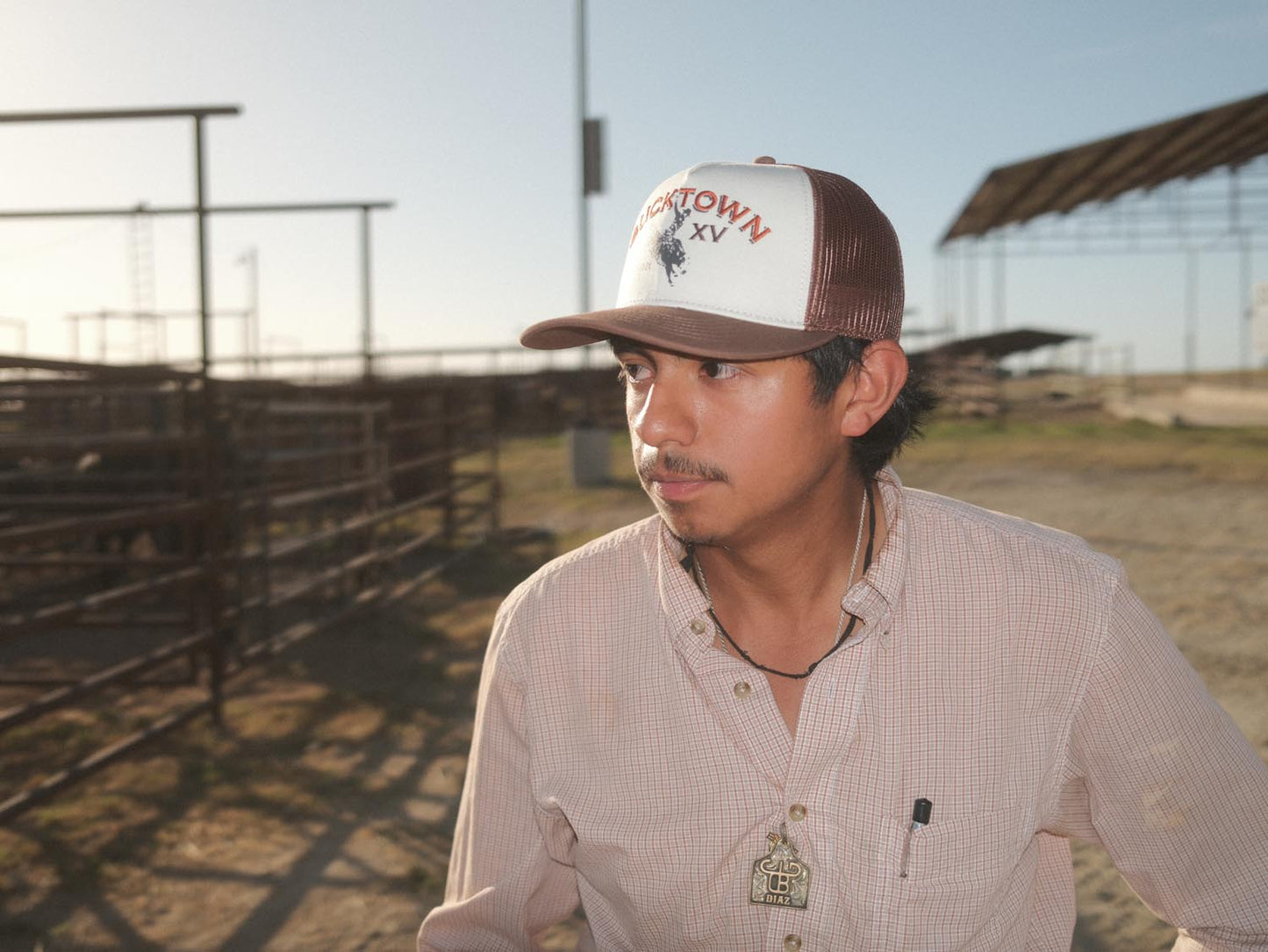 A man stands outdoors in a sunlit livestock pen area wearing the Rural Cloth Bucktown Original Hat, along with a plaid shirt and necklace, with metal fencing and open sheds in the background.