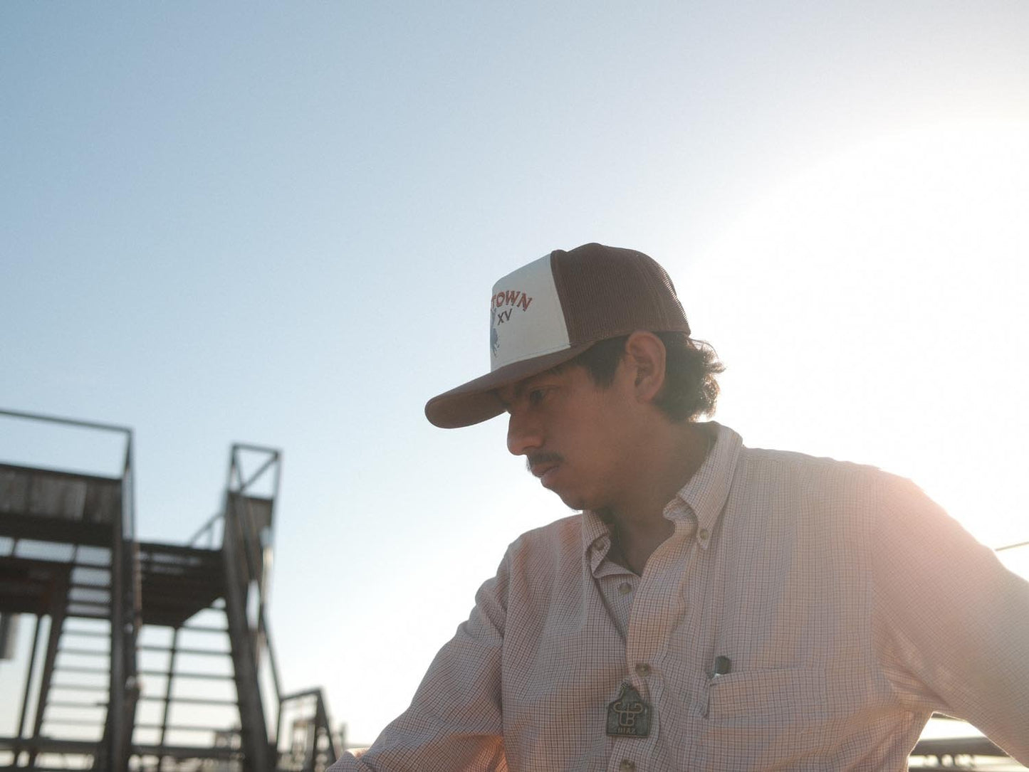A man outdoors by metal stairs, under a clear sky and bright sun, wears a Rural Cloth Bucktown Original Hat (adjustable snapback) and a button-up shirt.