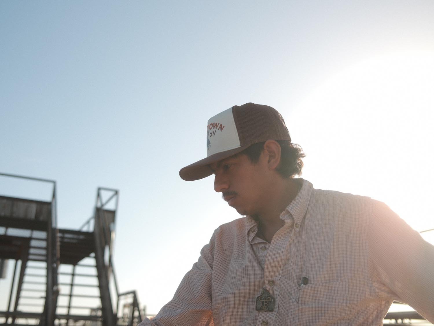 A man outdoors by metal stairs, under a clear sky and bright sun, wears a Rural Cloth Bucktown Original Hat (adjustable snapback) and a button-up shirt.