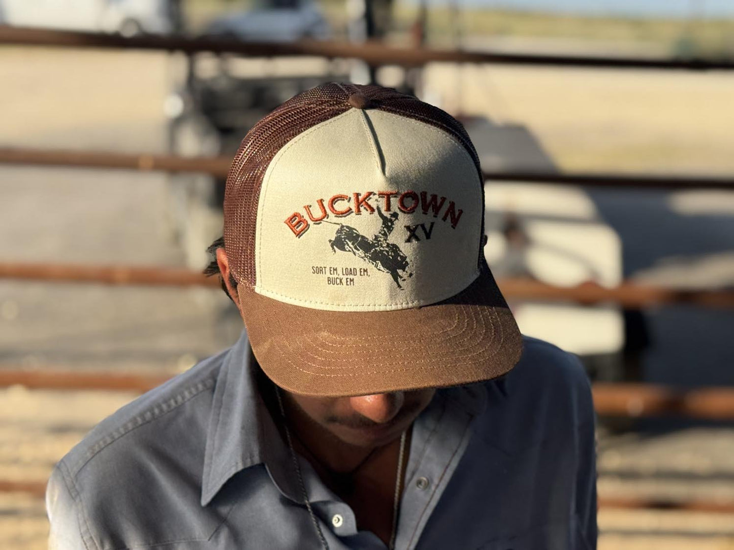 A person wearing a Rural Cloth Bucktown Original Hat with "BUCKTOWN XV" and a bull graphic on the front stands outdoors by a metal fence, their face partly shadowed.