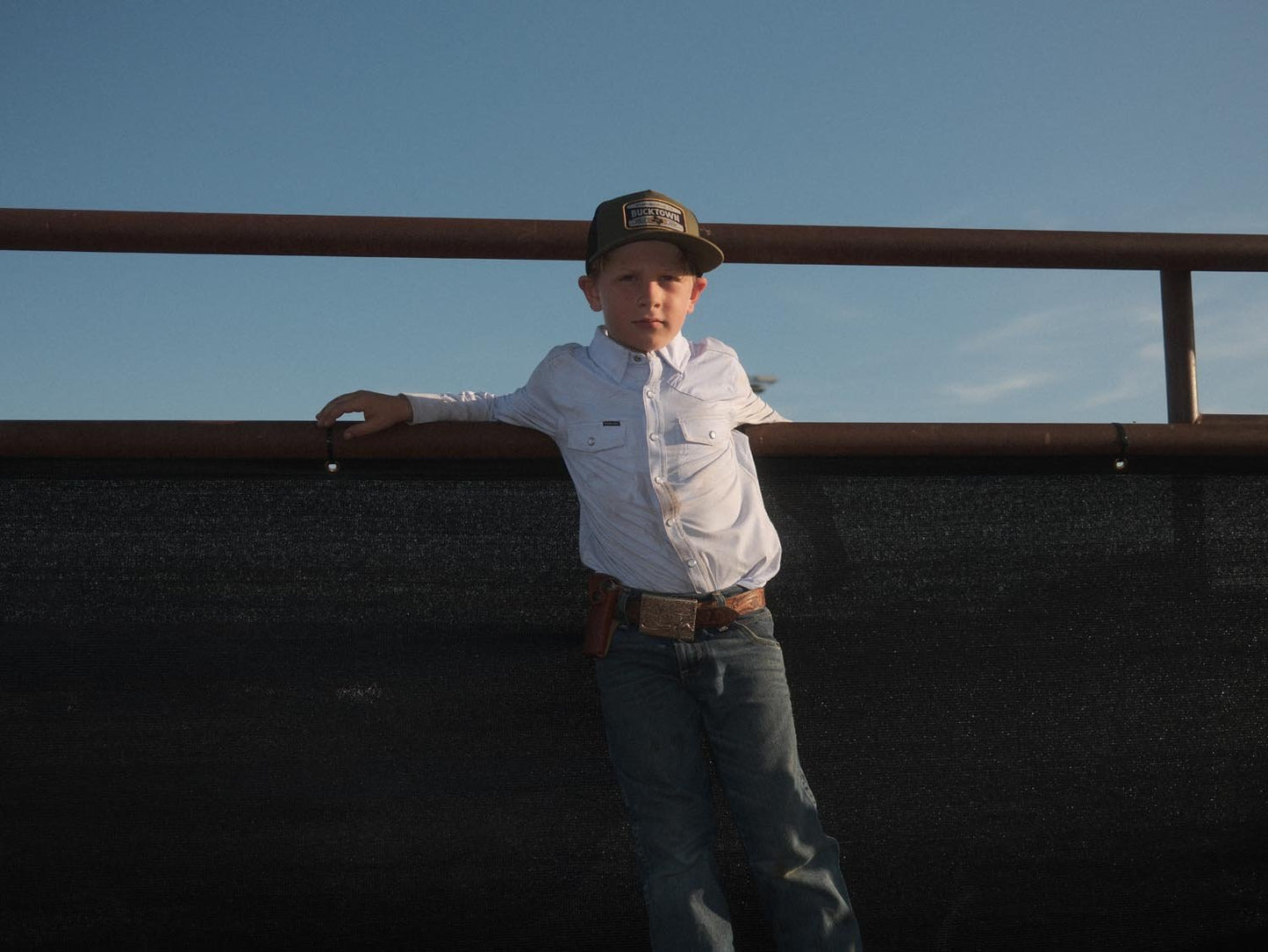 A young boy in a Rural Cloth Bucktown Outrider Hat, white long-sleeve shirt, and jeans stands by a metal fence at Bucktown XV Ranch, Stephenville TX, with one arm resting on the railing under a clear blue sky.