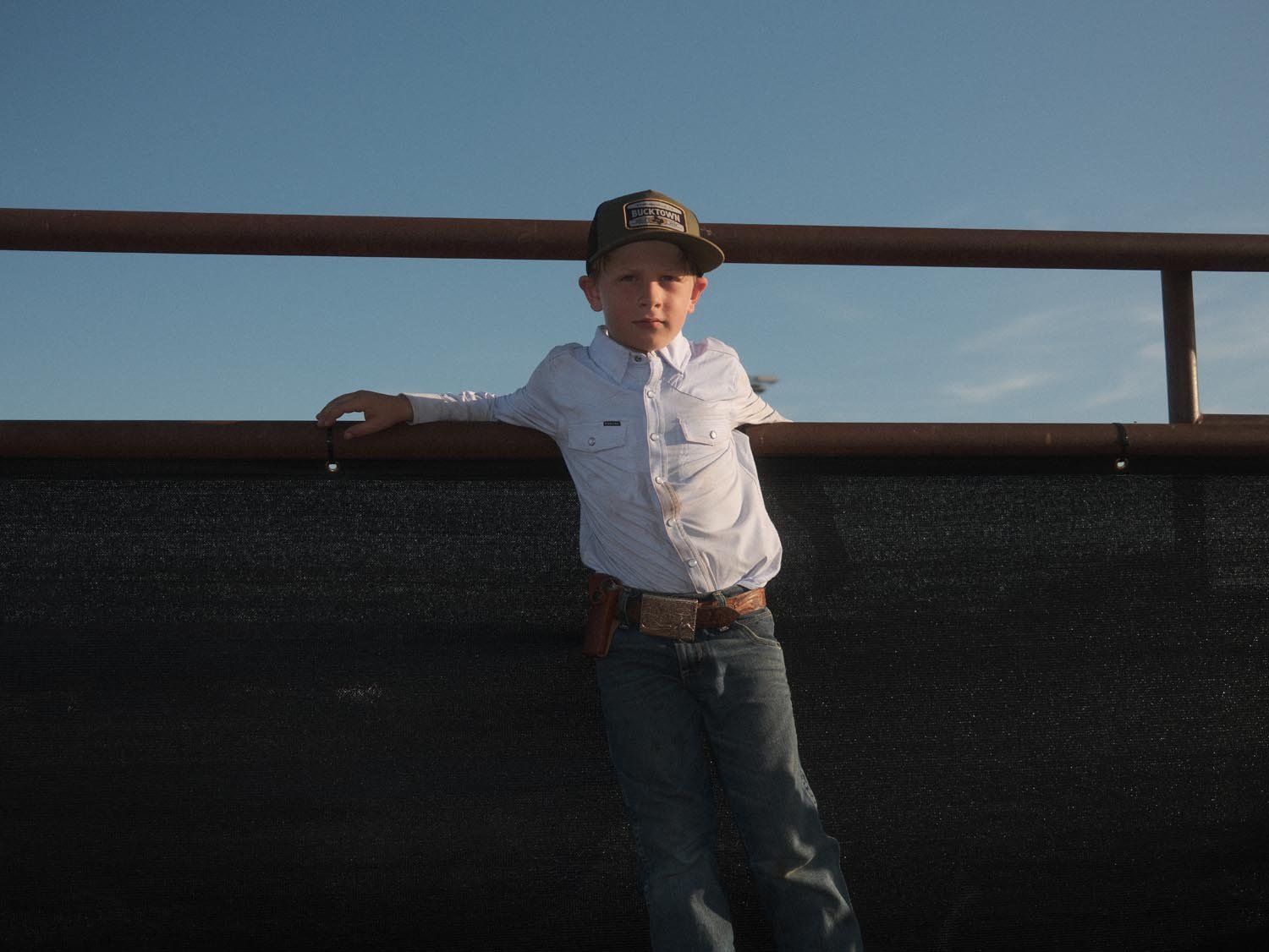 A young boy in a Rural Cloth Bucktown Outrider Hat, white long-sleeve shirt, and jeans stands by a metal fence at Bucktown XV Ranch, Stephenville TX, with one arm resting on the railing under a clear blue sky.