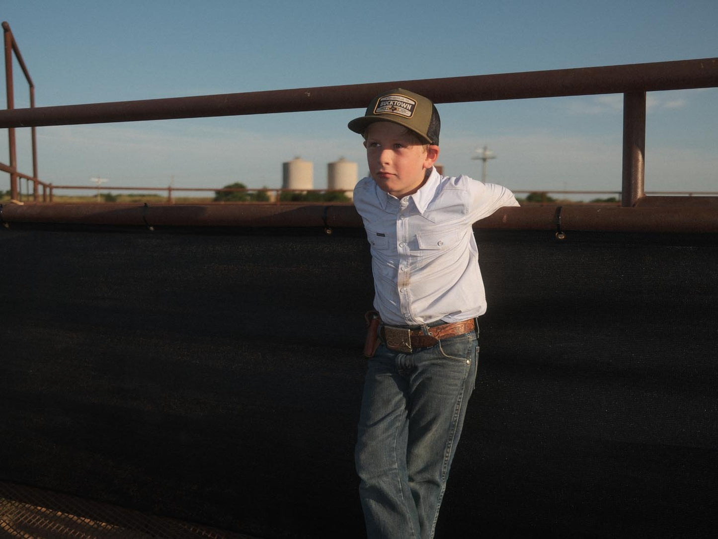 A young boy in a Rural Cloth Bucktown Outrider Hat, white button-up shirt, jeans, and a holster belt leans against a metal fence at Bucktown XV Ranch near Stephenville, TX, gazing off to the side under the open sky.