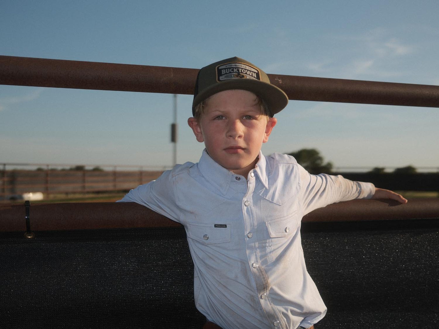 A young boy in a light shirt wears the Rural Cloth Bucktown Outrider Hat, standing by a metal fence at Bucktown XV Ranch near Stephenville, TX, with blue sky and open fields behind him.