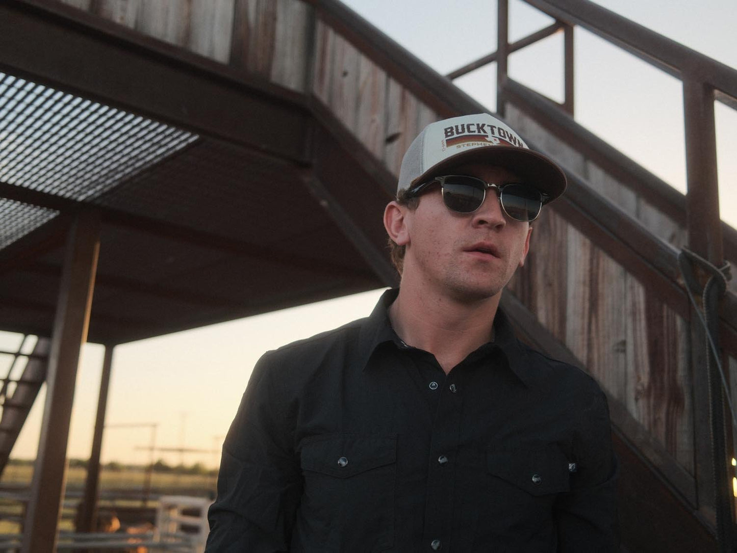 A man wearing a black button-up shirt, sunglasses, and the Bucktown Revival Hat by Rural Cloth stands before an outdoor staircase with a weathered metal structure at sunset.