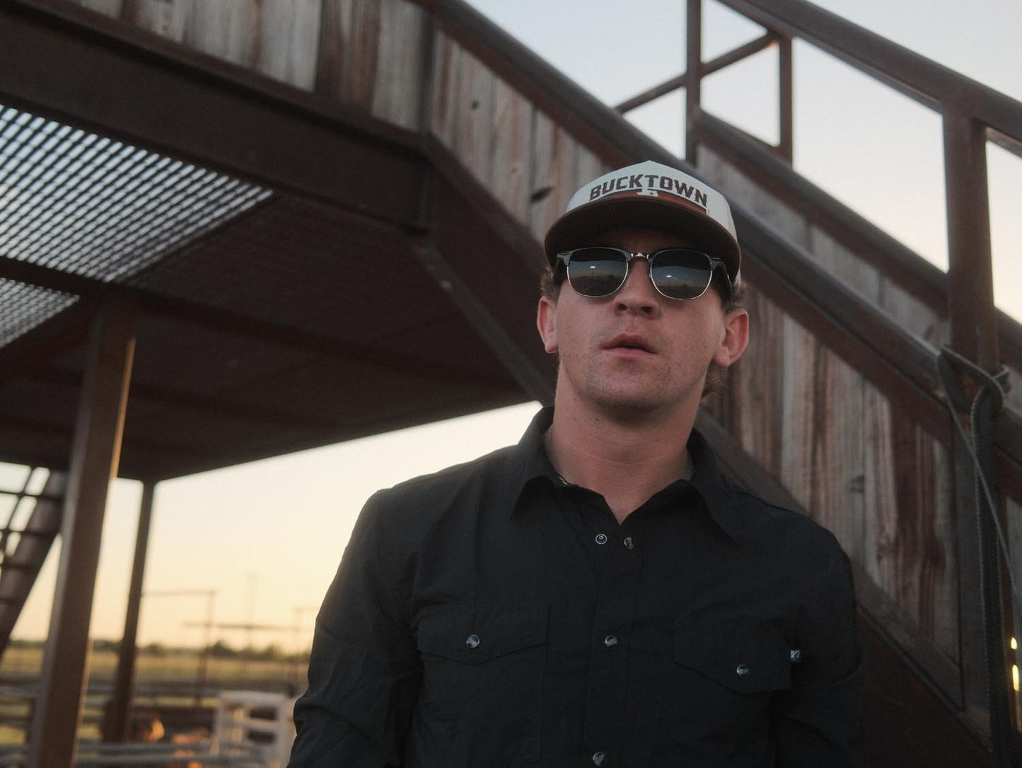 A man in a black button-up shirt and sunglasses wears the Bucktown Revival Hat by Rural Cloth, standing before a weathered metal staircase outdoors during daylight.