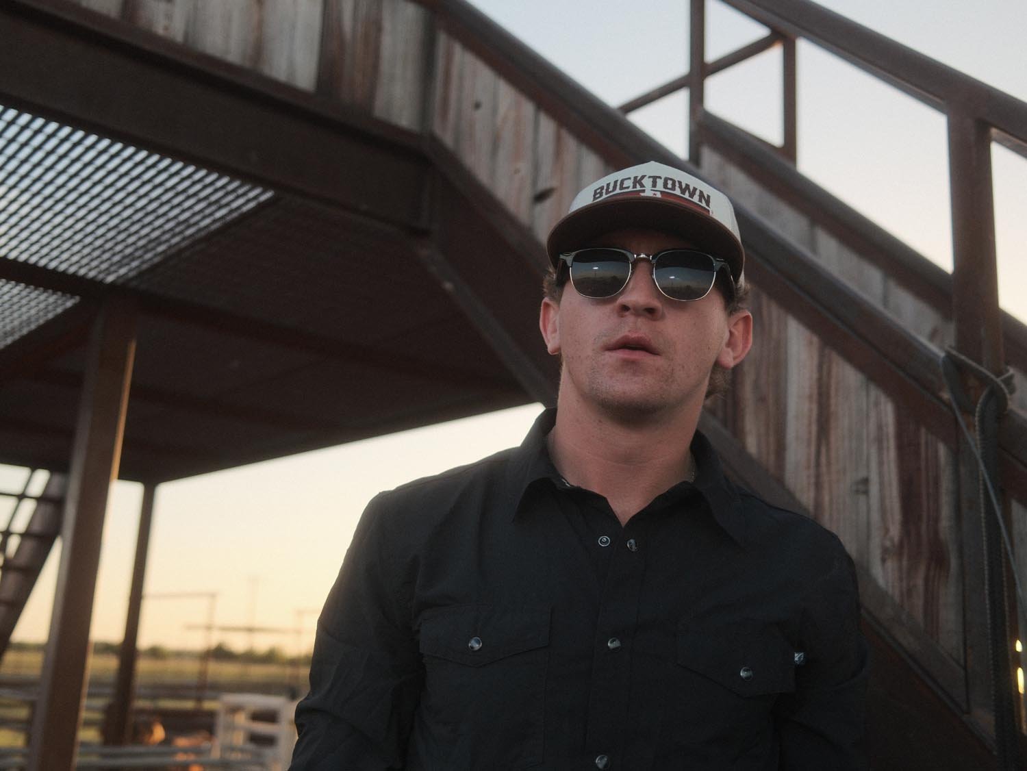 A man in a black button-up shirt and sunglasses wears the Bucktown Revival Hat by Rural Cloth, standing before a weathered metal staircase outdoors during daylight.