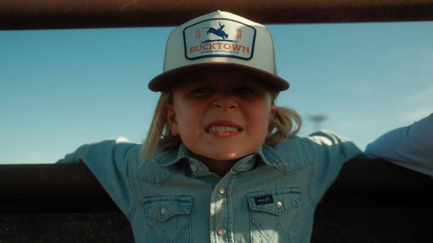 A child wearing a Rural Cloth Last Out Hat and a light denim shirt leans on a fence, smiling with arms outstretched under a clear blue sky.