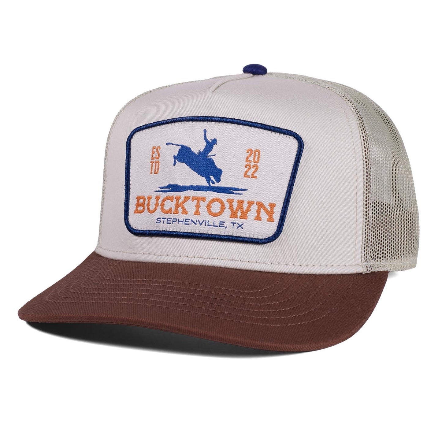 The Last Out Hat by Rural Cloth is a western snapback trucker with a white front, brown bill, beige mesh back, and a patch showing “BUCKTOWN STEPHENVILLE, TX ESTD 2022” plus an image of a person riding a bucking horse.