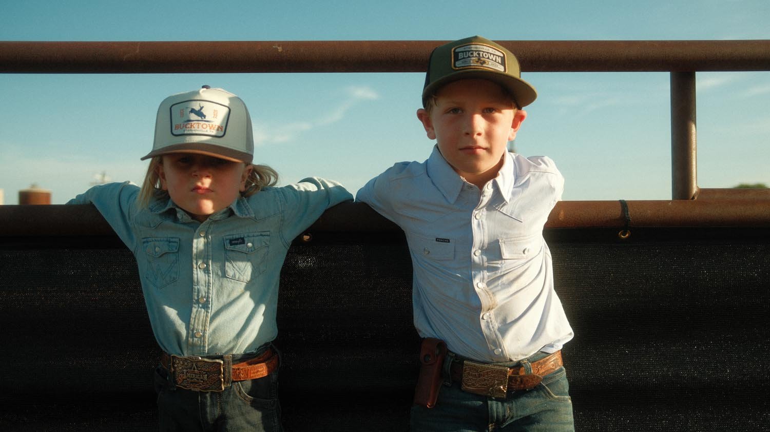 Two kids in button-up shirts, jeans, and Rural Cloth's Last Out Hat stand side by side by a metal fence under the blue sky at Bucktown XV Ranch.