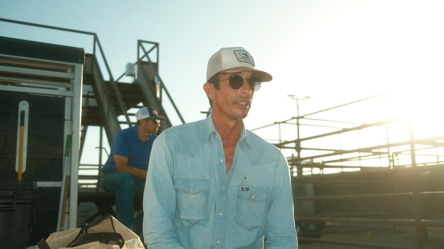 A man in sunglasses and a Rural Cloth Last Out Hat sits outdoors by a metal fence, dressed casually in a denim shirt. Another person in a cap and denim shirt is seated in the sunlit background.