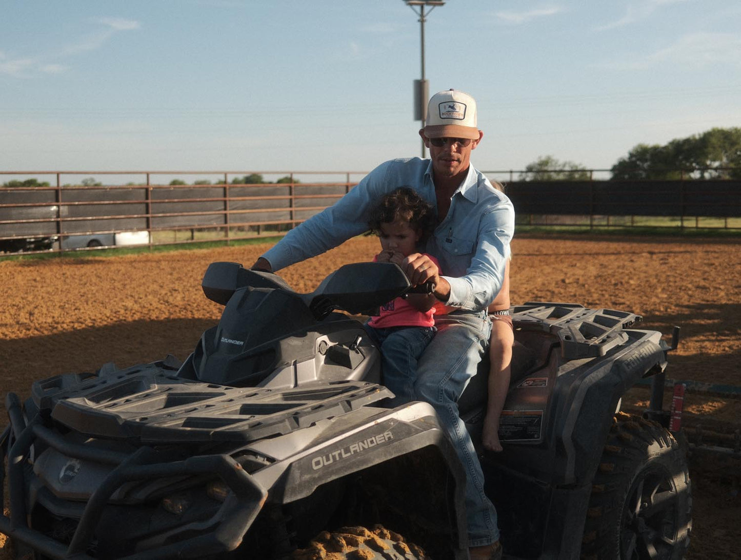 An adult in sunglasses and a Rural Cloth Last Out Hat sits on an ATV with a young child. The ATV is on dirt, with a fence and the open field of Bucktown XV Ranch in the background.