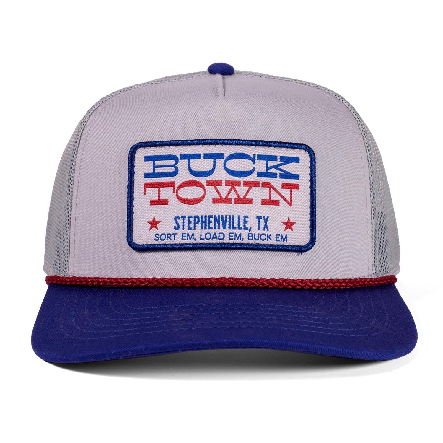 The Rural Cloth Patriot Hat is a gray and blue trucker hat with a red rope, featuring a "Buck Town" patch in blue and red. Below it reads "Stephenville, TX, Sort Em, Load Em, Buck Em" in red for an Americana look.
